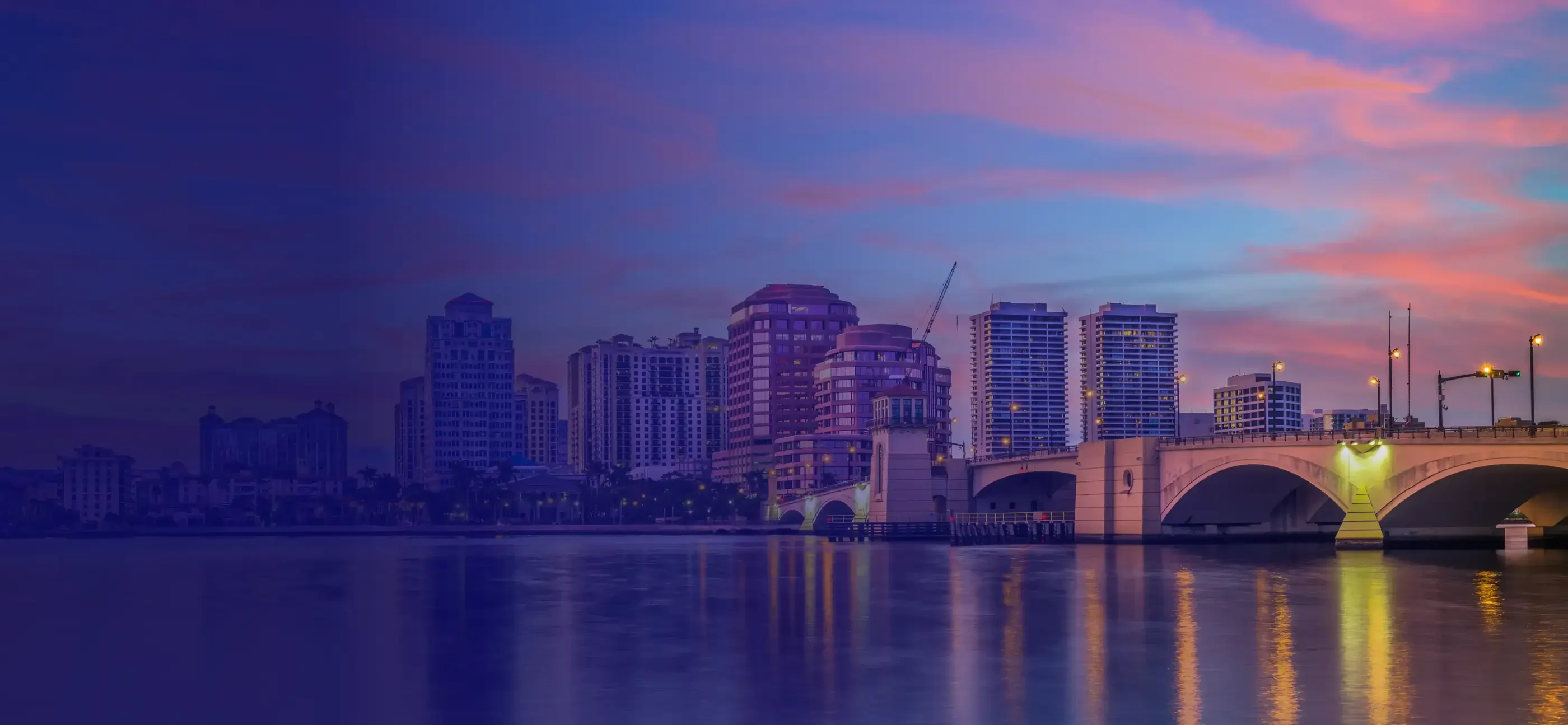 Skyline of a city at dusk, featuring modern buildings and a reflective waterway under a colorful sunset sky.