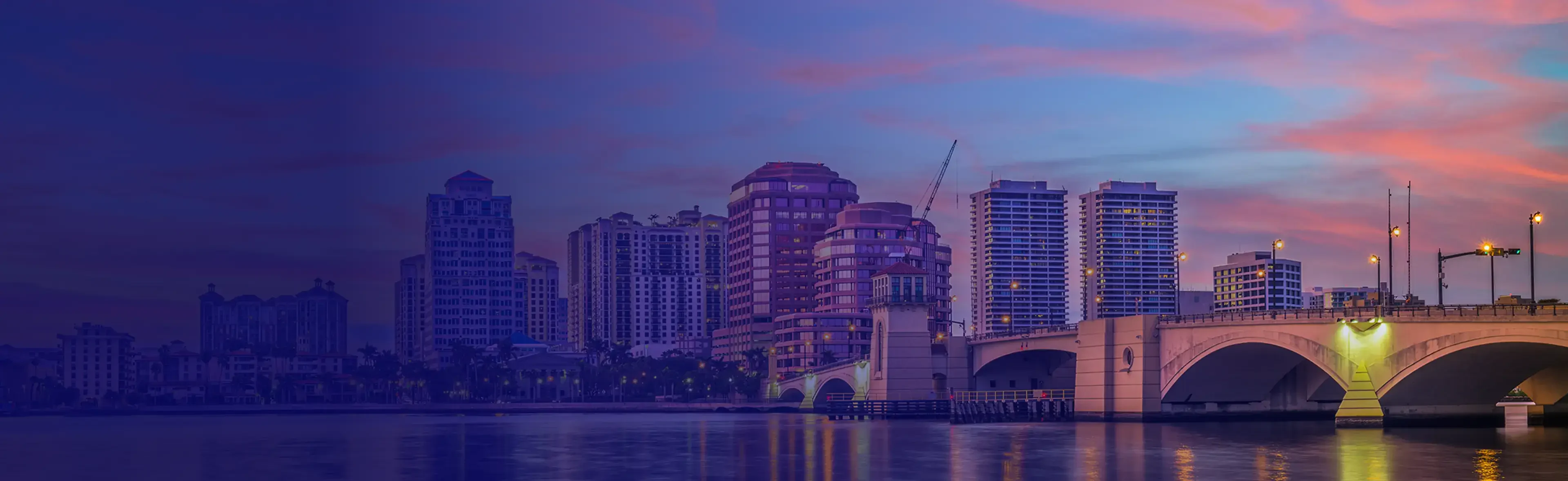 City skyline at dusk with modern buildings and a bridge reflecting in the calm water under a colorful sunset sky.